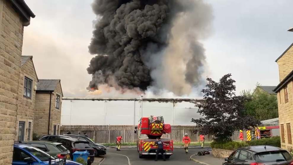 Thick black smoke rises in the air from the large grey industrial unit. A fire engine is parked at the end of Knoll Close which is a residential street of two-storey, almost identical beige brick buildings. There are lots of parked cars and three firefighters in hi-vis orange jackets, yellow trousers and white helmets are walking around the engine. A fourth firefighter in black overalls stands behind the engine which has a large ladder on it.