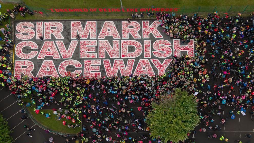 The mural from above. It is outlined in white with the names of children in red. There are hundreds of children standing around wearing bright colours.