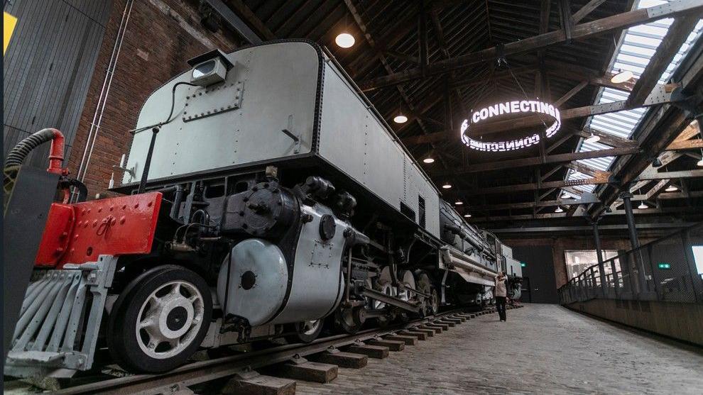 Inside the Power Hall is a large grey locomotive. A woman is staring up at the vehicle which is housed within the red-bricked warehouse. 