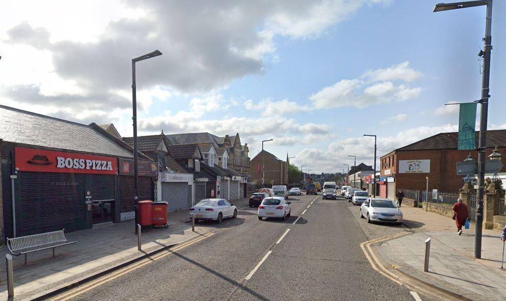 A Google Maps shot of a high street with cars, a pizza shop on the left hand side, a bench and pedestrians