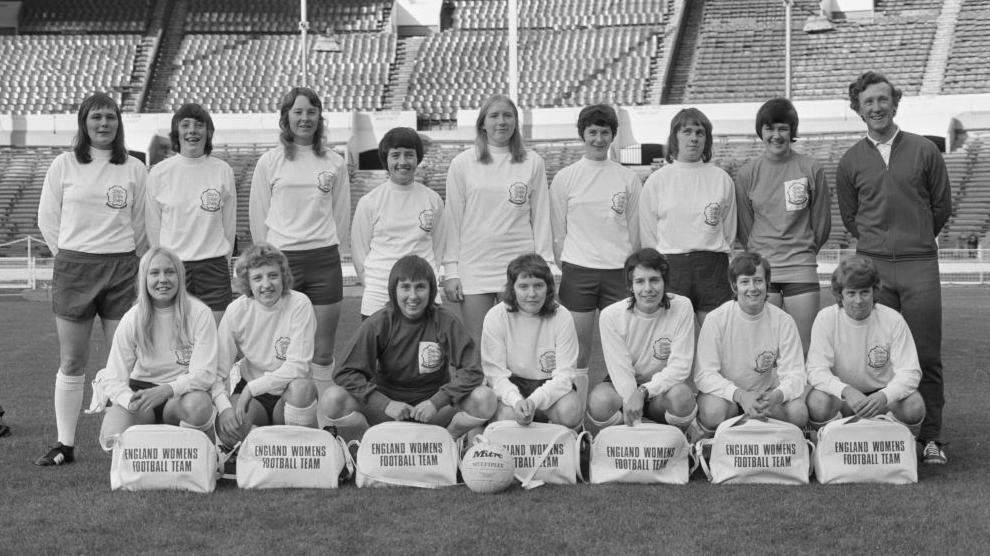 A black and white image of a group of women in football kit. They are stood in a football stadium.
