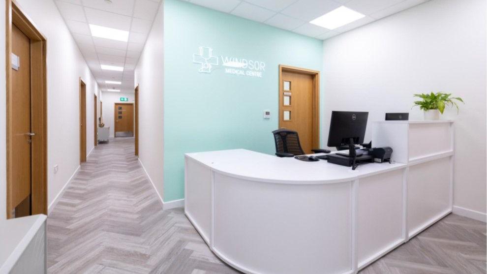 A medical office reception area. The reception desk is white and curved, with a computer and a potted plant on it. The reception is empty. The room is white painted with a feature wall in a duck egg colour with the company logo on it in white writing. To the left of the reception area is a hallway with several wooden doors.