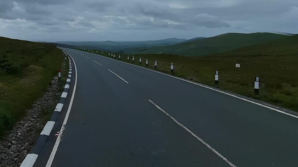 The Mountain Road just beyond the Brandywell junction, which has a grass embankment to the left and hills to the right.