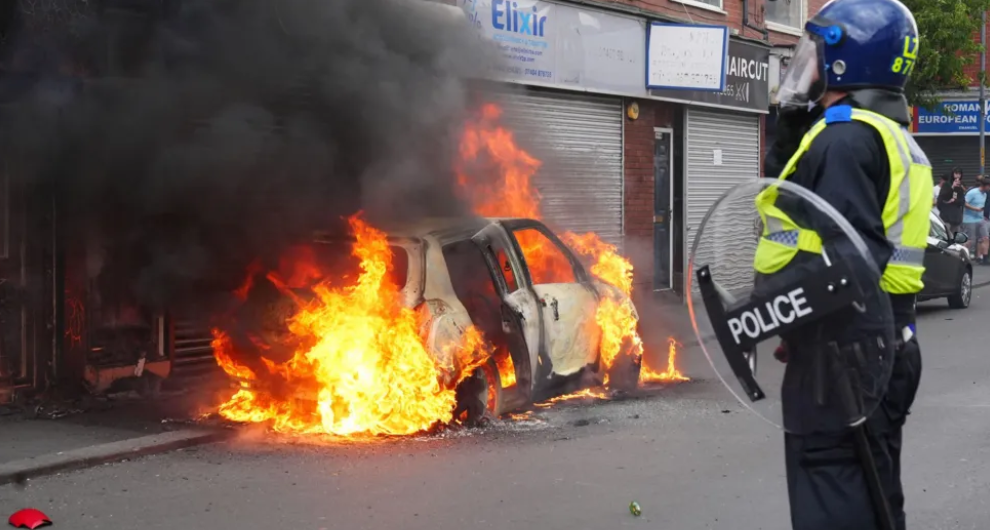 A police officer in riot gear standing in front of a car on fire in front of shuttered shops.