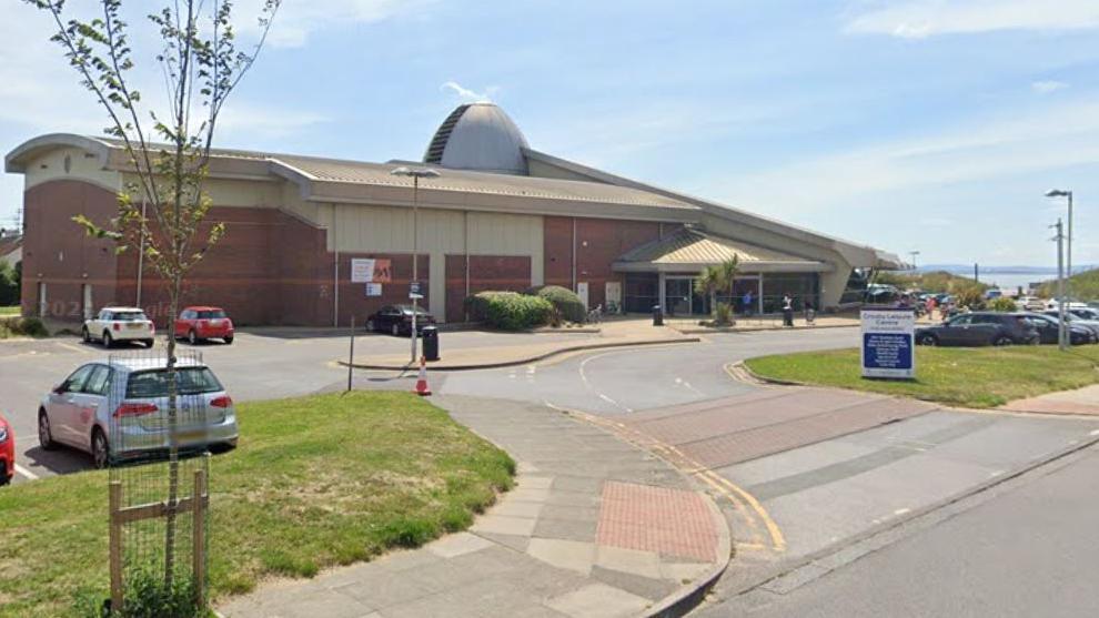 A red and yellow brick building with a dome top and large glass entrance. It has a large car park with a sweeping entrance and several cars parked. There are sand dunes in the background.