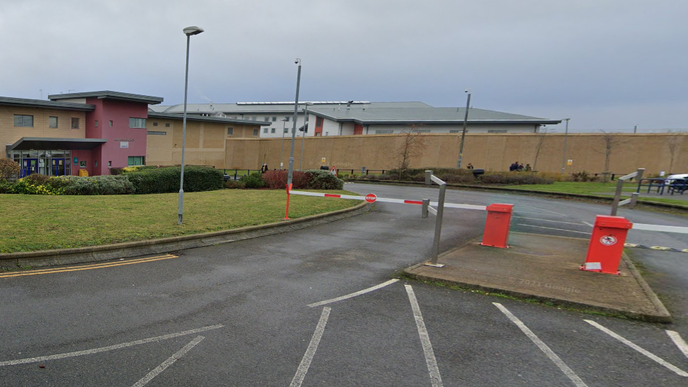 The entrance to a modern prison. There are rising barriers across a road leading to the entrance block, which looks like a two-storey office block. Beyond a high, solid wall is a higher prison building.