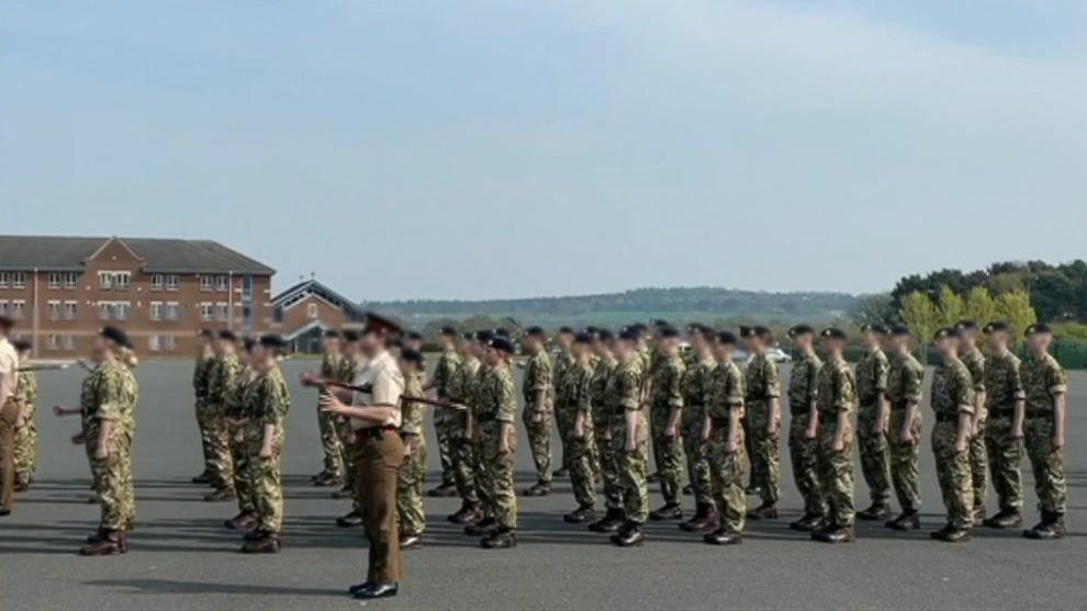 A screenshot from a video showing a regiment practicing their uniform marching and saluting on camp at Larkhill. There is a large group of about 30 people dressed in green and brown camouflage uniforms and black caps. They are standing on a large tarmac area surrounded by fields and trees. In the background there is a brown brick building with lots of windows.