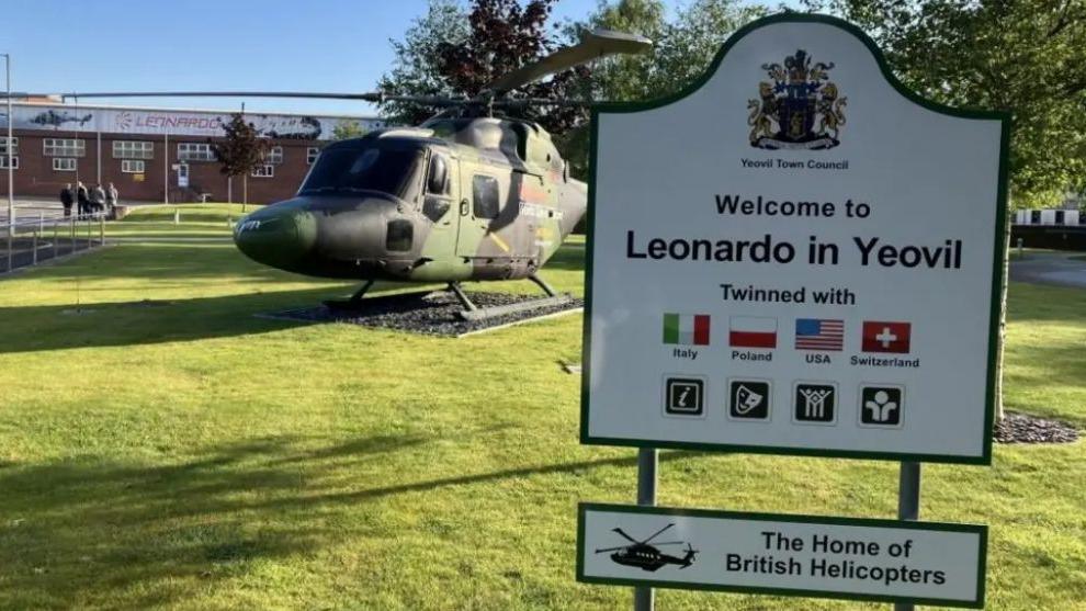 The outside of Leonardo factory in Yeovil. In the foreground, a sign reads "Welcome to Leonardo in Yeovil" and another below reads: "The Home of British Helicopters". In the background, a camo helicopter can be seen on lawn, with the factory behind it.