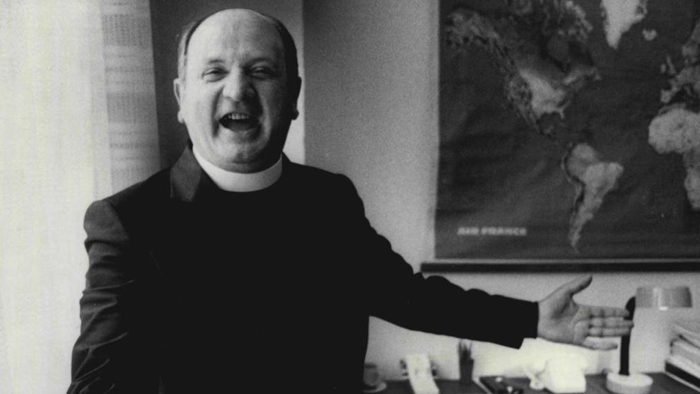 A black and white image of Bishop Eamonn Casey, wearing a black suit and smiling. There is a map of the world on the wall behind him.