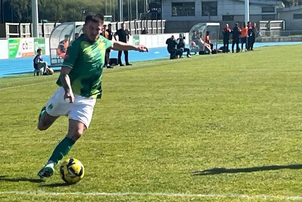 A grassy football pitch on a sunny day. A footballer with short dark hair in a green top, white shorts and green socks pulls his leg back to cross the ball, while running at pace.