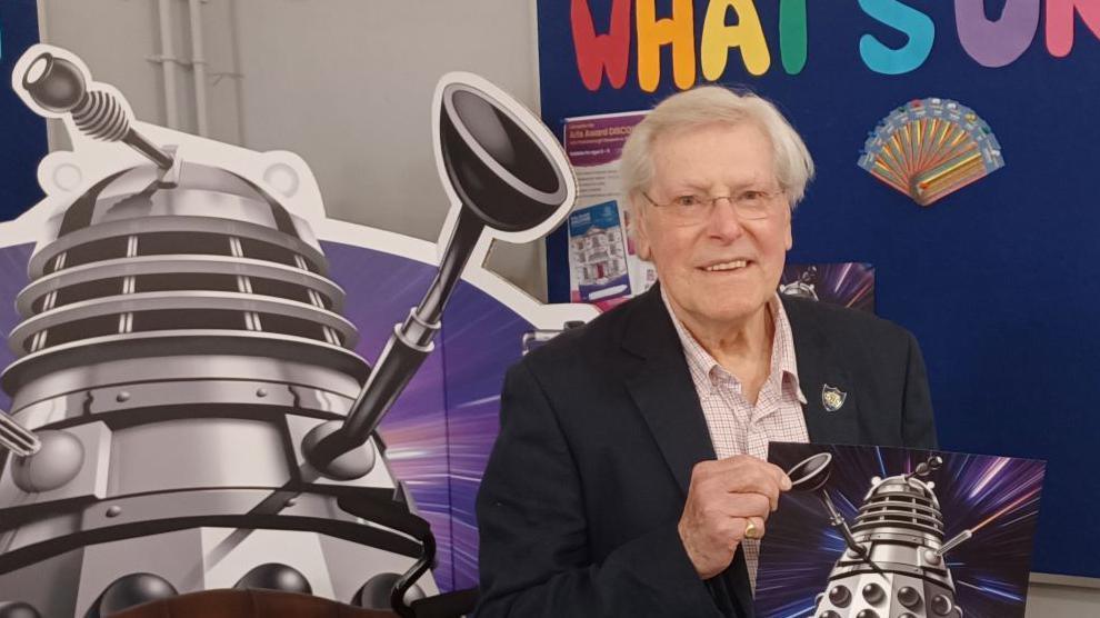 Peter Purves, a white-haired man in his eighties, wearing glasses, a dark blue jacket, white checked shirt and light brown trousers, stands behind a desk, holding up a poster reading ‘Adventures in Space and Time’ and displaying an image of a Dalek, a conical robotic creature. Behind him is a large cardboard cut-out of a Dalek.