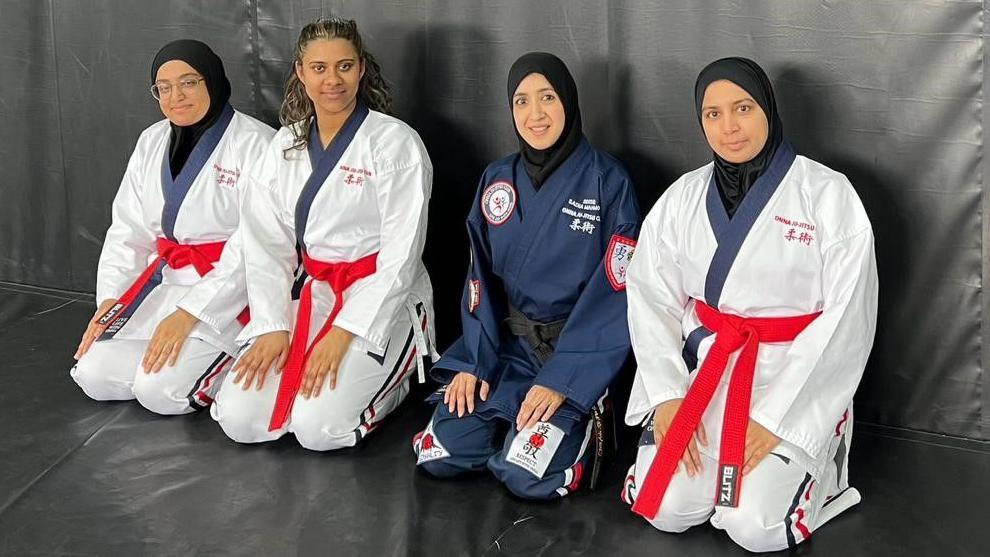 Four women sat in a row wearing martial arts uniforms, three women have white suits with red belts and one woman has a dark blue suit and black belt