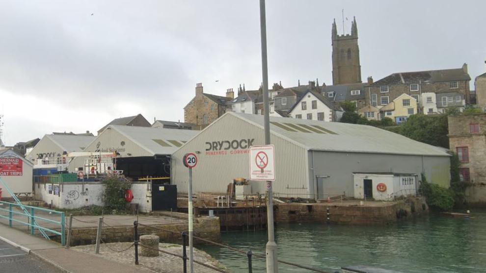 Penzance's dry dock. The water level sits just below the building. Above the water level are three conjoined grey buildings. They have the word "drydock" written on them. Behind the dry dock is a row of houses and a church steeple.