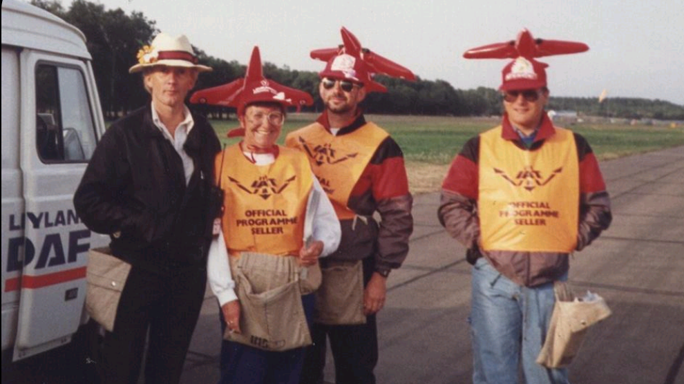 Three people are wearing planes as hats while volunteering to sell RIAT posters, standing on a runway. A fourth person, a man wearing a hat, is standing with his hands in his coat pockets looking at the camera.