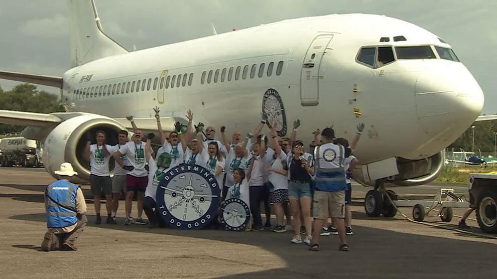 A group of people cheering in front of an airplane, near the right wing. They are holding two wheels with an inscription "DETERMINED TO DO GOOD". A man in a white hat and blue top in front of them is on one knee, appearing to be taking a picture.