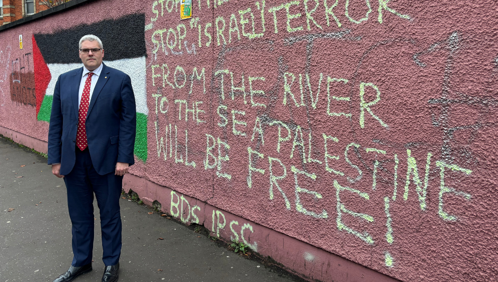 DUP leader Gavin Robinson stands to the left of a wall, which has been daubed with the phrase “from the river to the sea, Palestine will be free”