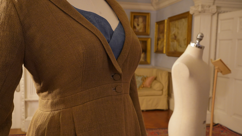 Close-up of the top of a linen-style v-necked overcoat with two buttons on a mannequin. It is in a room from a stately home with gold-framed pictures on the walls.