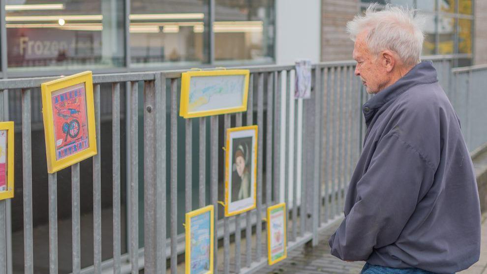 A man with grey hair, wearing a blue jacket looking at a number of yellow framed art pieces that are attached to a metal fence.