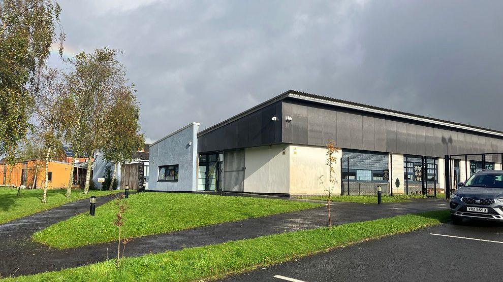 Exterior view of Bunscoil an tSléibhe Dhuibh in west Belfast.   It is a modern building surrounded by some trees, a lawn and paved walkways.  A car is parked in its carpark.