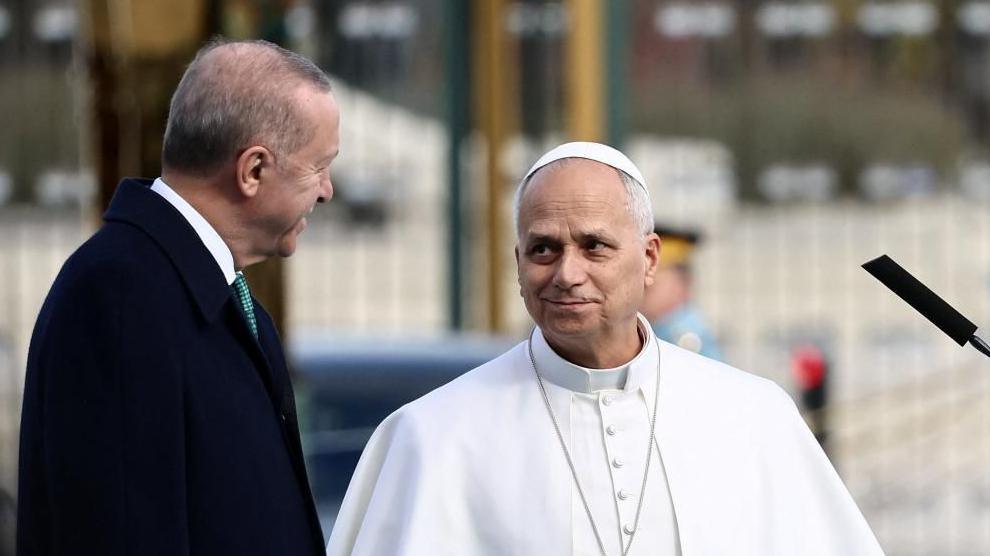A man in white clerical garb talks to a man in a black business coat. 