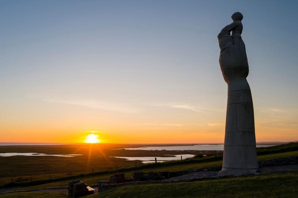 The statue, which depicts the Virgin Mary with baby Jesus in her arms, overlooks moorland and the sea. The sun is glowing on the horizon.