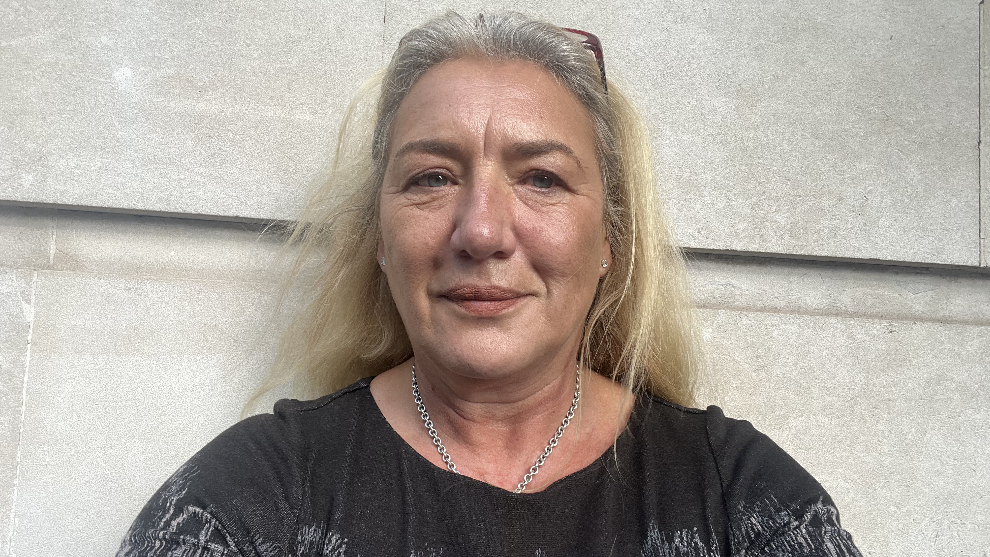 Melissa Moore stands outside her office against a wall, wearing a dark top, silver necklace, with glasses holding her blonde hair in place