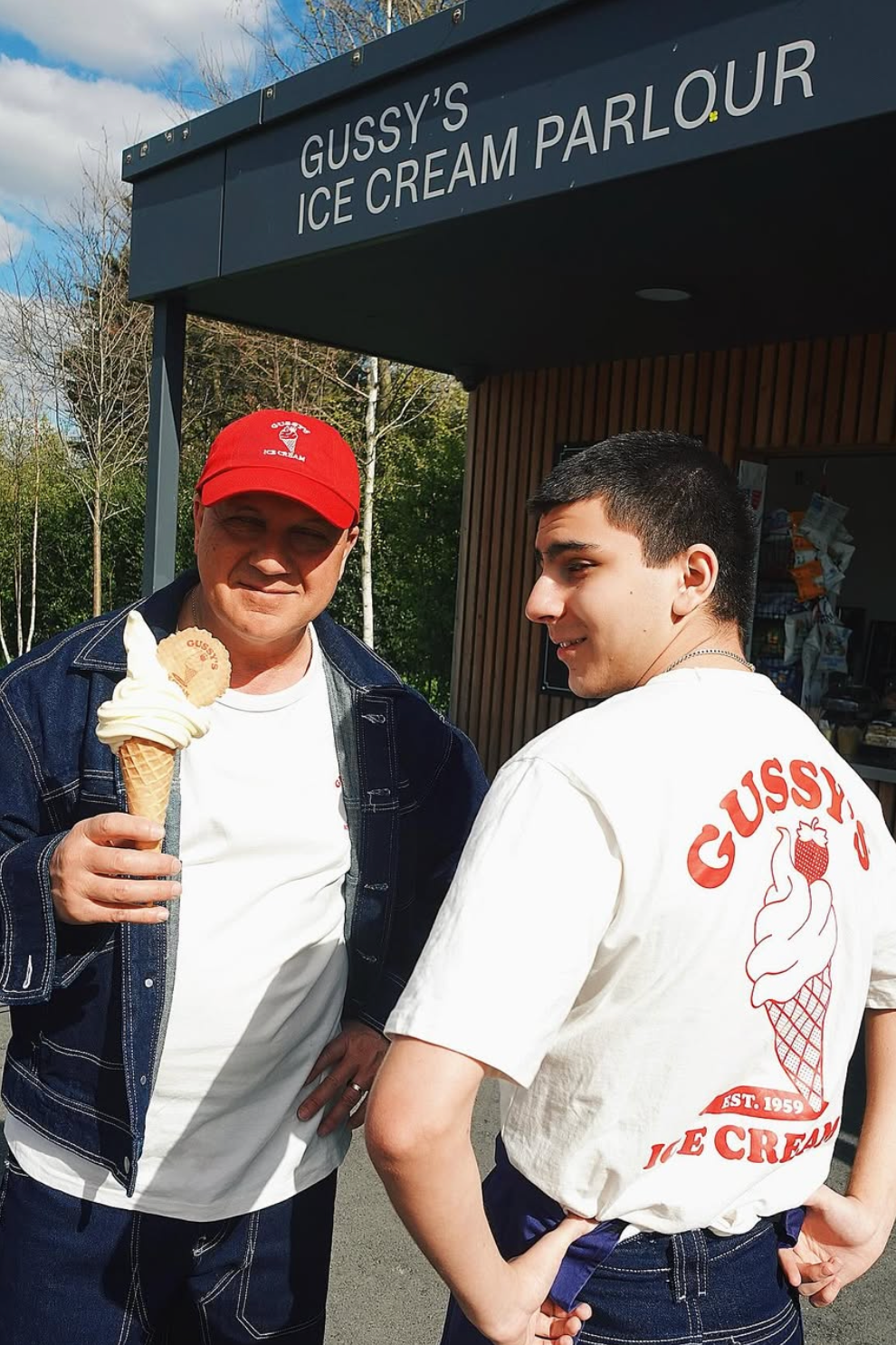 Two people stand outside Gussy’s Ice Cream Parlour. Gus holds a soft-serve ice cream cone, while his son Oli wears a white T-shirt featuring a large red ice cream graphic and the words “Gussy’s Ice Cream Est. 1959” on the back.