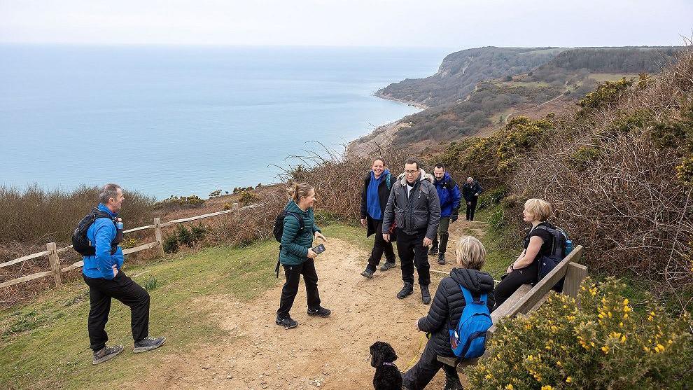 A group of walkers are gathered, some standing, some sat on a wooden bench, high above the sea from Hastings to Fairlight Cove. A small black dog sits next to one of the walkers sat on the bench.