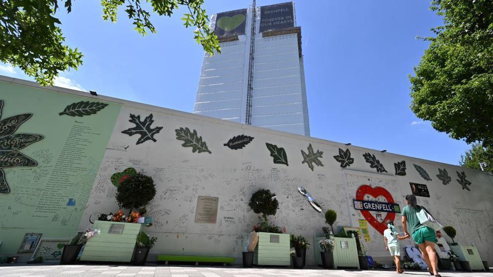 A woman and girl look up at Grenfell Tower from in front of a wall covered in written tributes