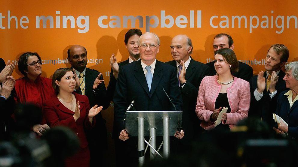 Sir Menzies Campbell is standing at a podium with microphones. He is wearing glasses and a dark suite, white shirt and light blue tie.He is surrounded by his supporters who are clapping and smiling at him. There is an orange backdrop with the words ming campbell campaign