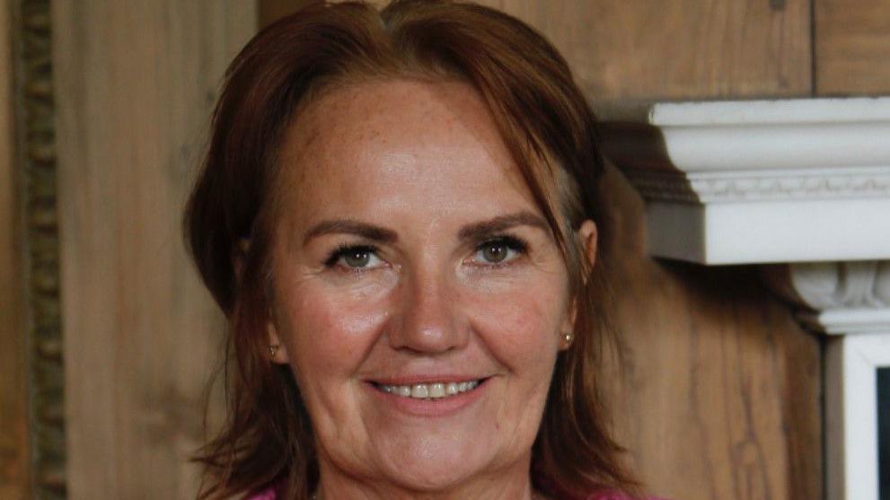 A head shot photo of a woman looking into the camera.  She's smiling.  She has shoulder length brown hair and bluey-grey coloured eyes.  