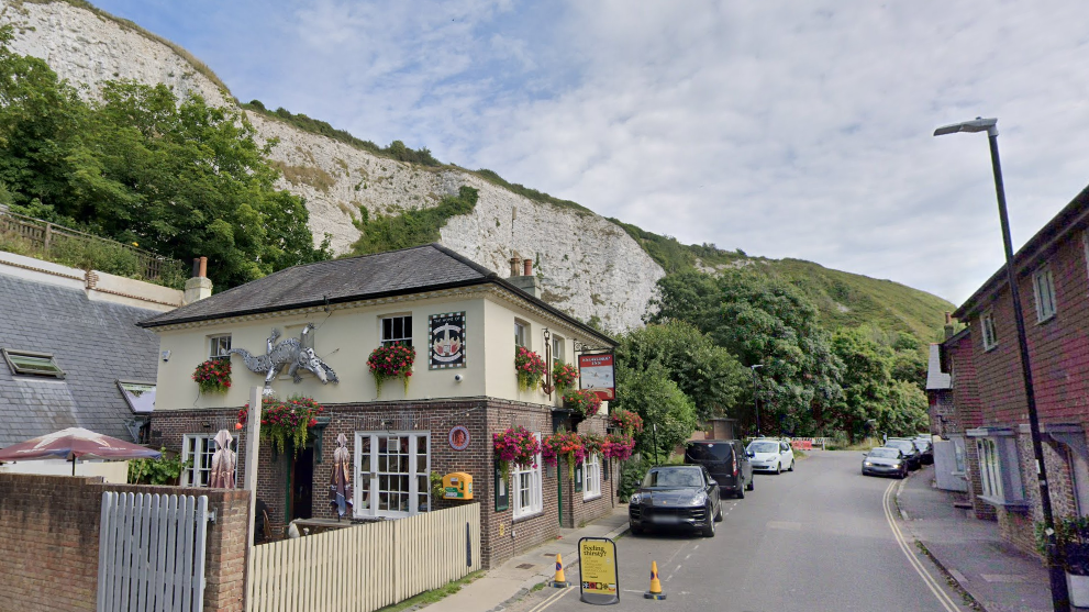 A old fashioned pub beside a narrow road. The pub and road are beneath white cliffs which are covered in trees and grass