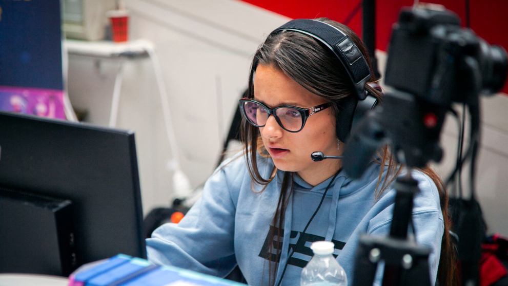Shows a girl in a blue hoodie, glasses and headset with mic playing a computer game