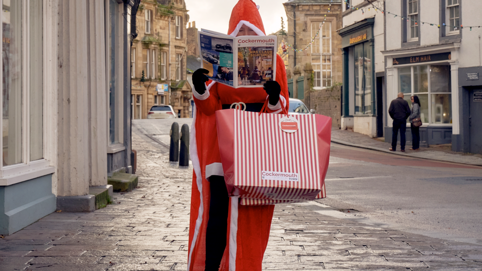 A woman dressed in a red Santa suit, holding a newspaper and a shopping bag, standing on a cobbled street. 