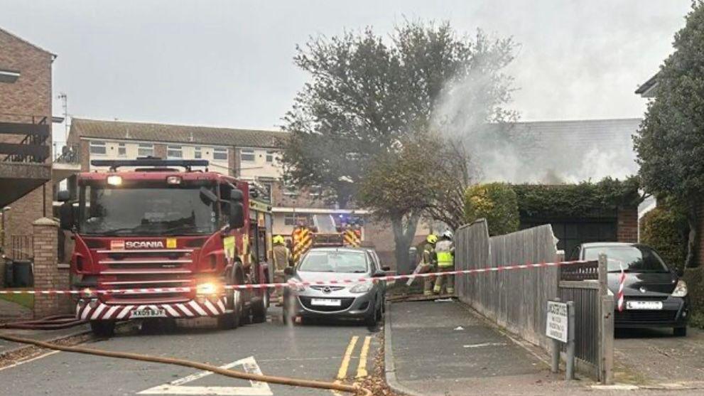 Smoke rises from behind a fence and hedge, with a fire engine and two cars parked in front of them. Two fire fighters are looking over the fence and the scene is sealed off with red and white incident tape.
