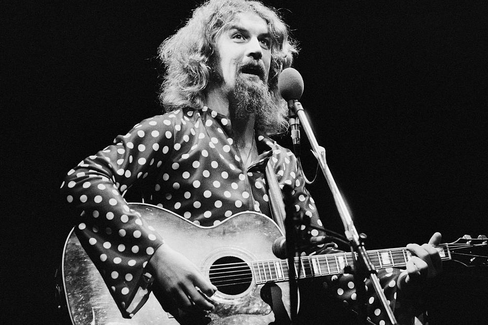 Black and white photo of Billy Connolly. He is playing a guitar and singing or talking into a mic. He has long hair and a long beard and is wearing a spotty shirt