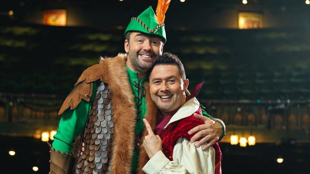Jason Manford and Ben Nickless in pantomime costume stood on stage smiling at the camera. Nickless is pointing at Manford.