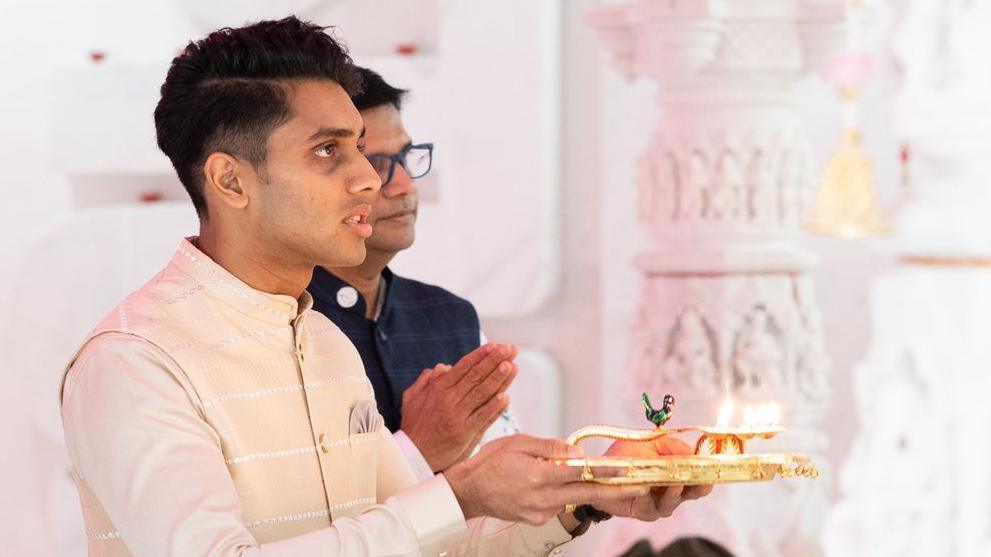 Dev is in a cream traditional Indian top performing a prayer. He holds is a gold coloured plate with a diva. 