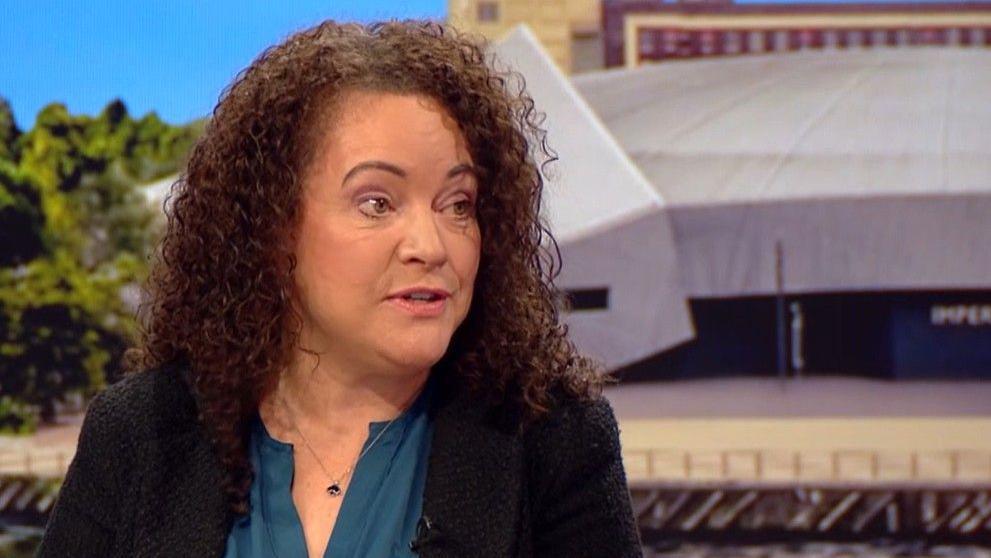 Rachel McMillan has long permed auburn hair a turquoise blouse and dark blue jacket.