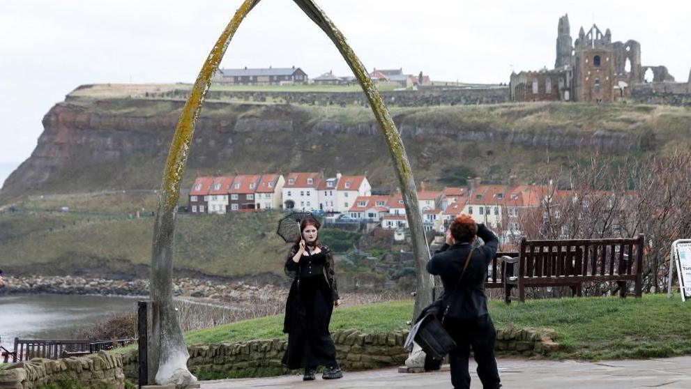 A person dressed in black Victorian-style clothing holding a parasol stands beneath a large arch made from whale jawbones on a coastal promenade. Another individual is taking a photo of the scene. In the background, there are red-roofed houses on a hillside and the ruins of Whitby Abbey overlooking the town and the sea.
