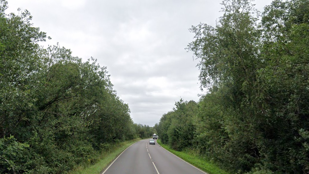 Two cars are driving along the A1122, there are green hedges either side of the road.