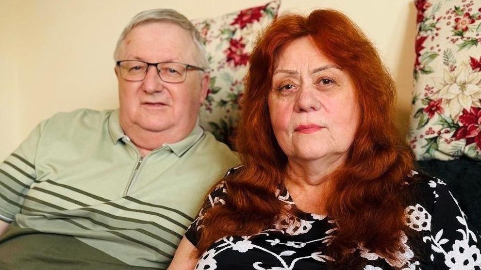 Man and woman - Anatolii and Lidiia Kyrylov - looking at camera, the man with grey hair and glasses, wearing a green top, and a woman with red/brown hair and a black and white top.