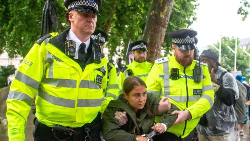Shows a woman in a green coat being carried away in handcuffs by two police men in high viz yellow vests during the pro-Palestine Action demonstration in London