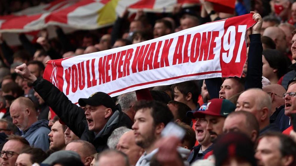 A crowd of Liverpool Football Club supporters holding a banner which reads 'You'll Never Walk Alone 97' 