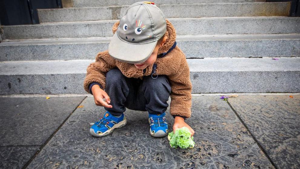 A boy holding a dinosaur toy and wearing a hat with cartoon eyes on it looks down on the paving slab with the fossils