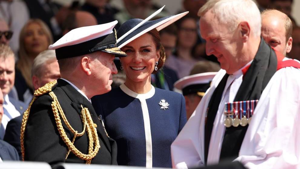 The Duchess of Rothesay wearing a navy dress with white binding, and matching hat. She had a thistle broach and long earrings. She is smiling, and standing alongside a man in military uniform and another man in priest like dress. The Duke of Rothesay is slightly visible in the corner.