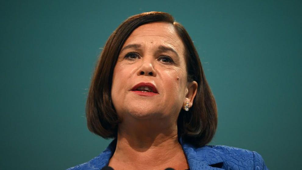 Mary Lou McDonald is talking and looking at something above the camera line. She has dark shoulder-length hair and is wearing a blue blazer. The background is solid green. 