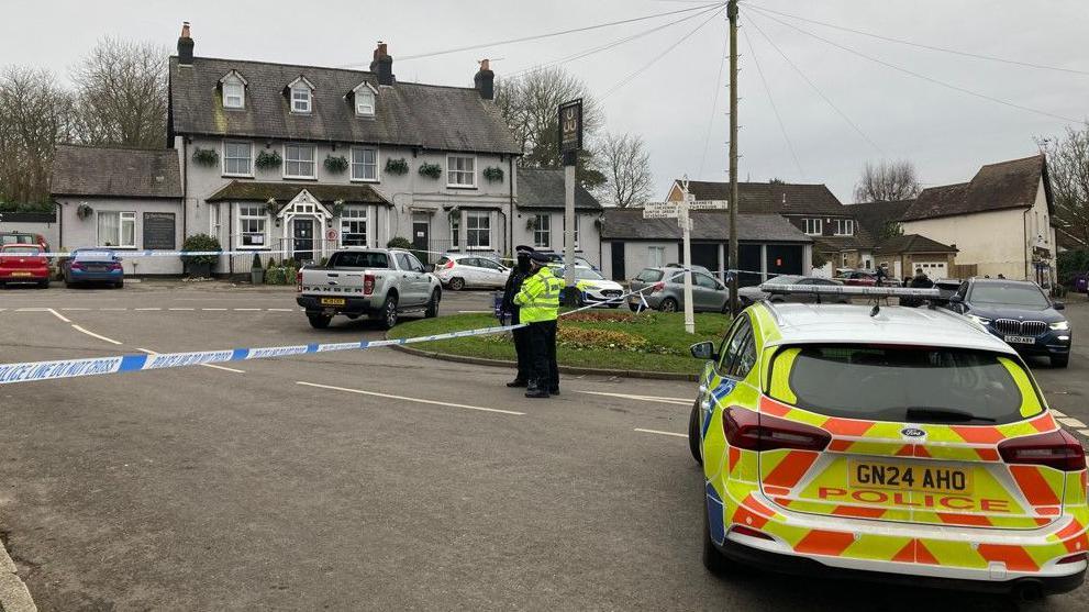 Police stand outside the pub, which is taped off. A police car is in the foreground.