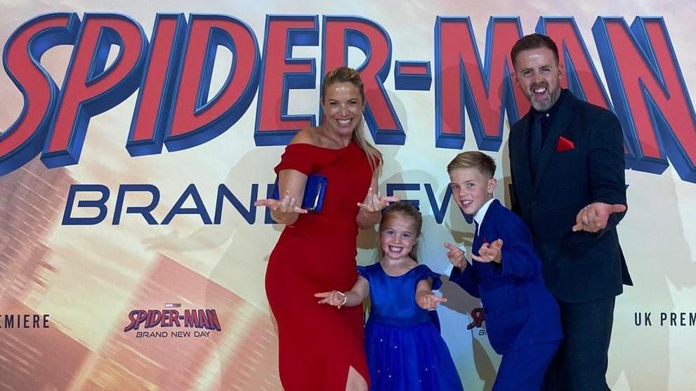 A blond woman in a red dress, man in a black suit, and two children in royal blue do the spider man pose, extending their hands to the camera, in front of the premiere poster.