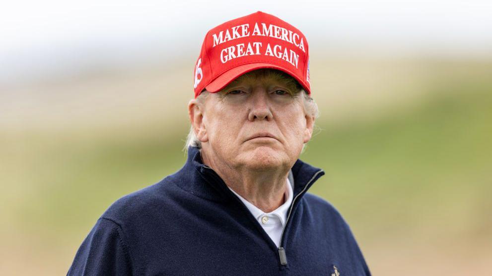 US President Donald Trump with Turnberry course in the background. He is wearing a red Make America Great Again cap.